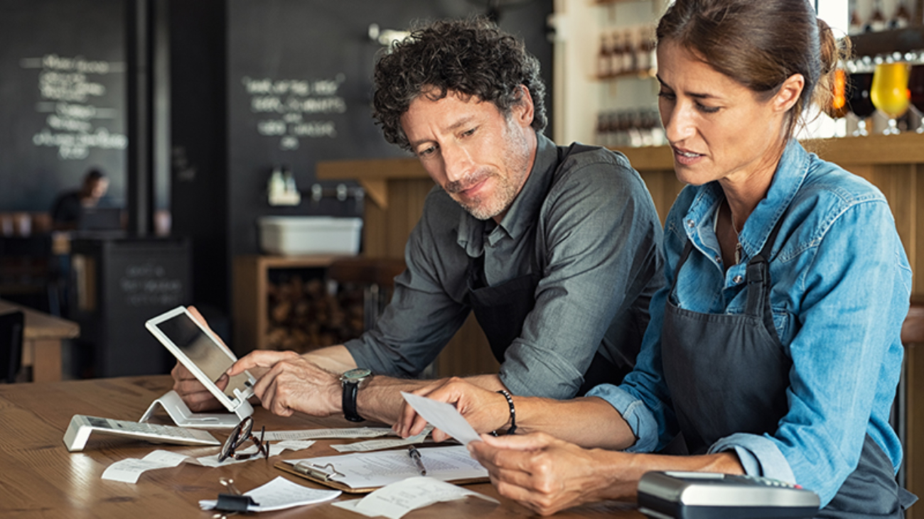 Man and woman sitting in cafeteria discussing finance for the month. Stressed couple looking at bills sitting in restaurant wearing uniform apron. Café staff sitting together looking at expenses and bills.